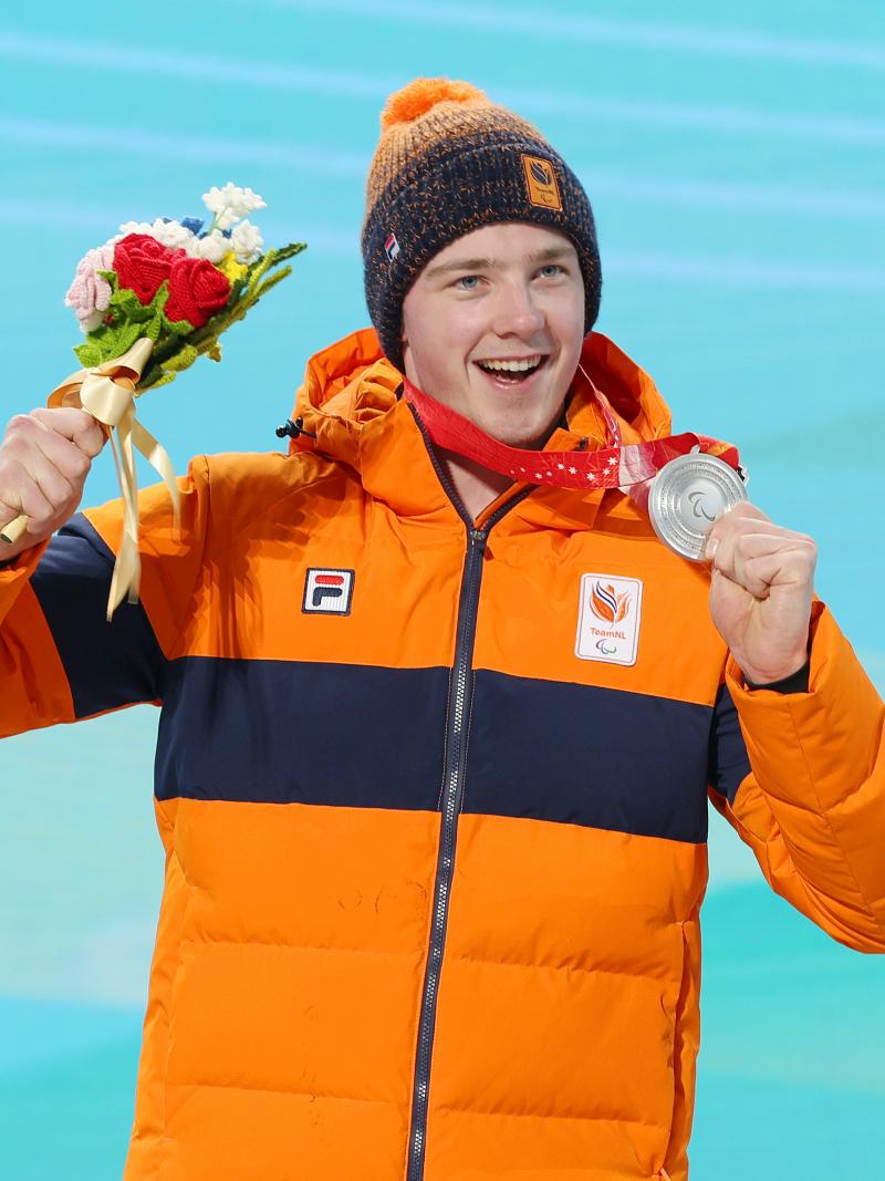 A smiling male athlete holds up a silver medal and bouquet of flowers during a medal ceremony. 